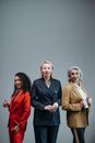 Home Three diverse professional women confidently posing in elegant business suits with a neutral gray background in a studio setting.